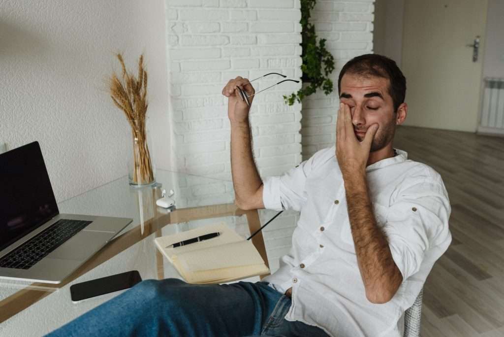 Ultra Bann A tired man in a white shirt rests at a home office desk, holding eyeglasses and rubbing his eyes